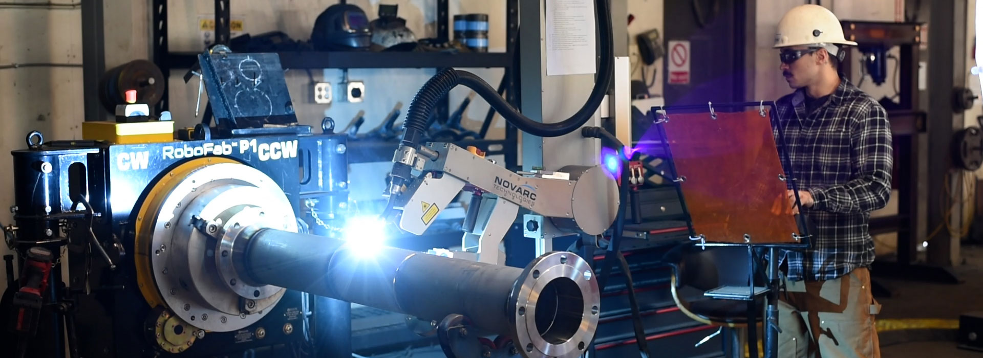 A worker in a hard hat and safety glasses operates a robotic welding arm on a large metal pipe in an industrial workshop. Shelves with tools and equipment are visible in the background.